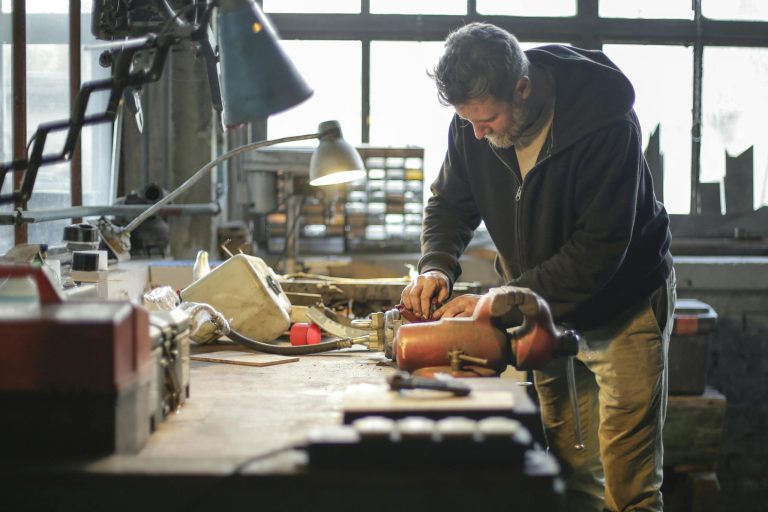 Side view of serious middle aged male in workwear standing near workbench and working with tools and metal details in lamplight in workshop